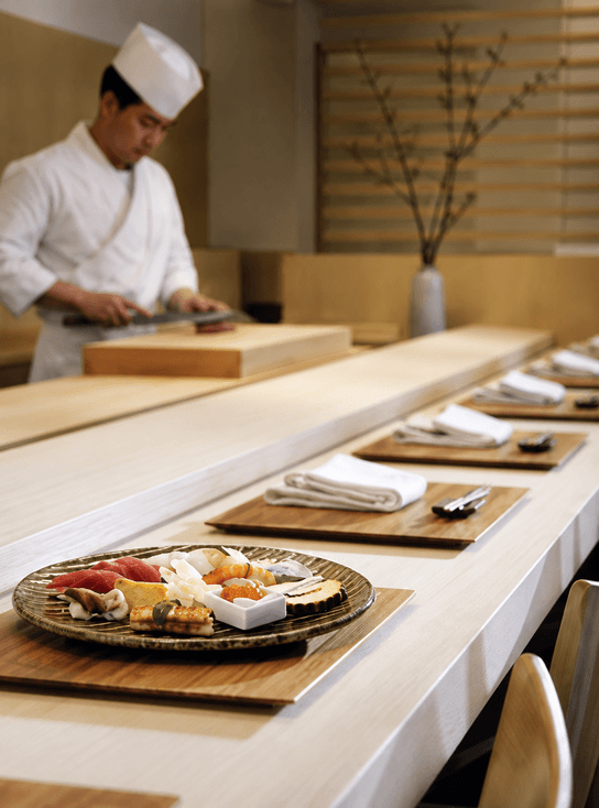 A counter set with plates of sashimi and side dishes before a chef at work in a quiet dining room.