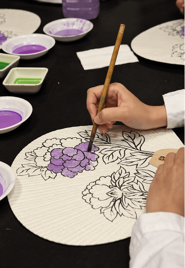 A brush painting a peony in purple on a porcelain plate.